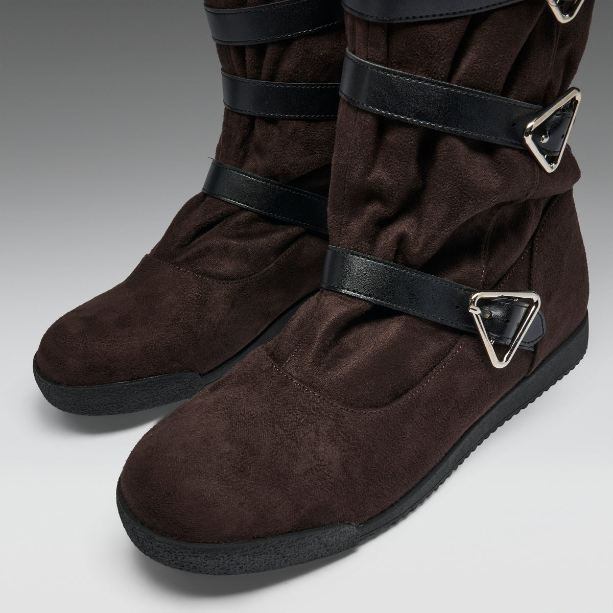 A pair of dark brown suede boots with black straps and silver triangular buckle details, shown on a neutral gray background.