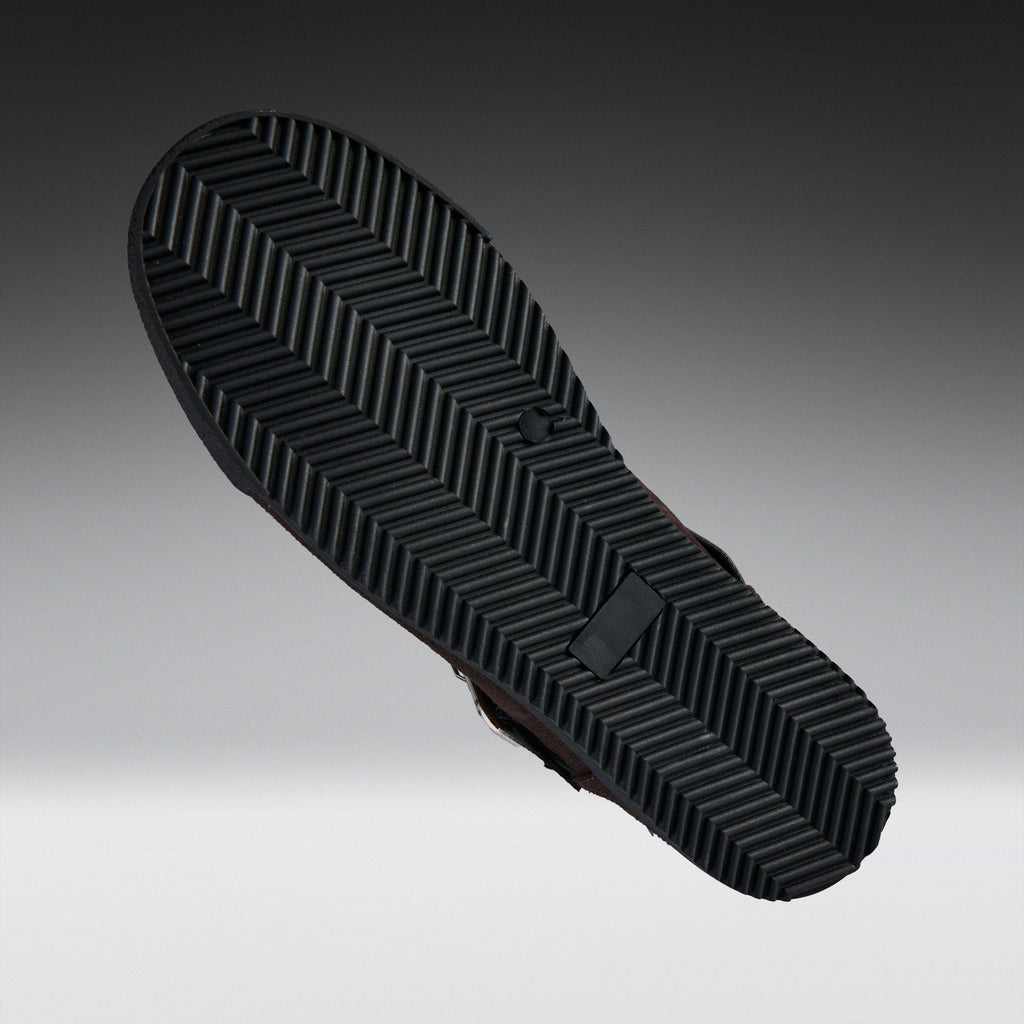 A close-up of a black shoe sole with a textured chevron tread pattern, shown against a gradient gray background.