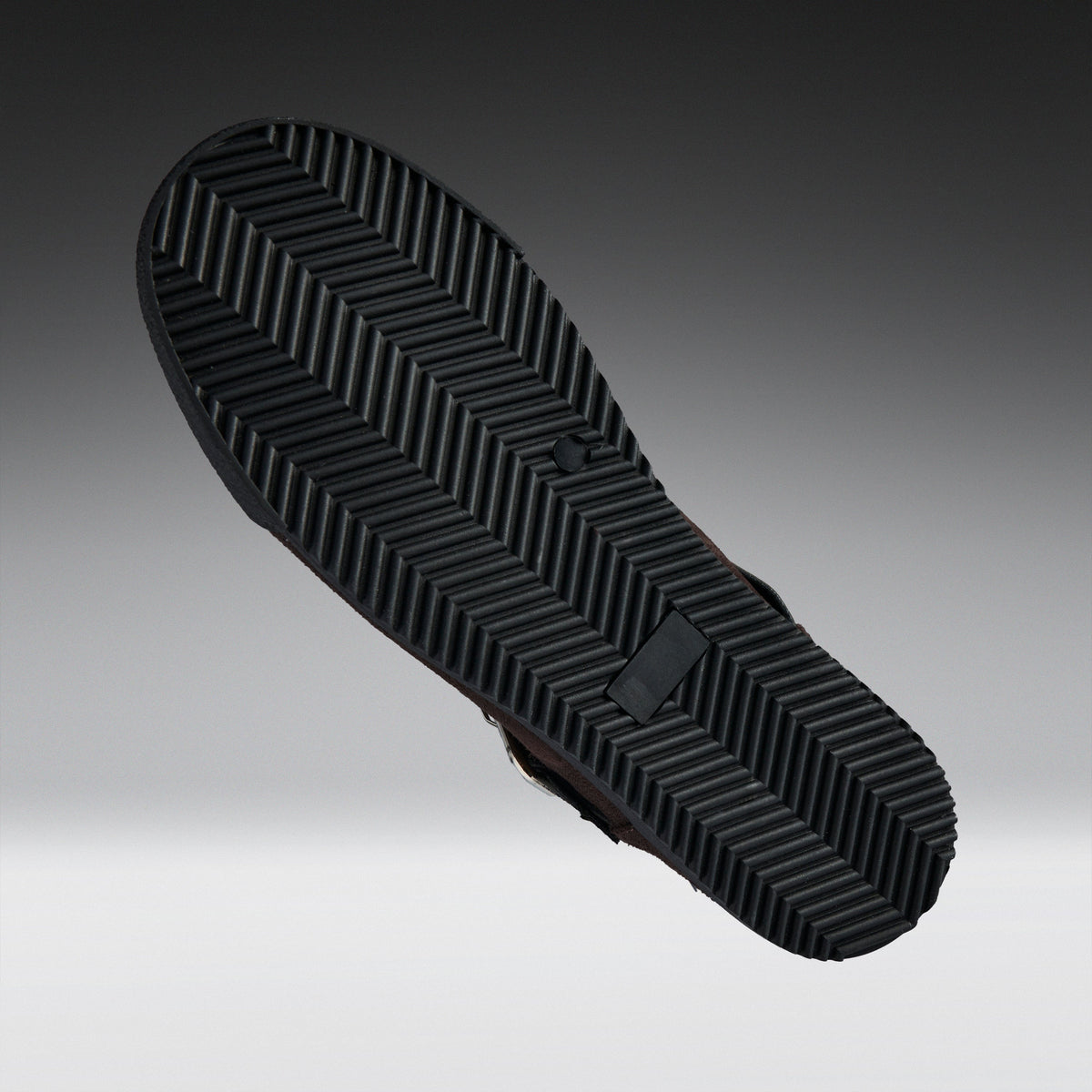 A close-up of a black shoe sole with a textured chevron tread pattern, shown against a gradient gray background.