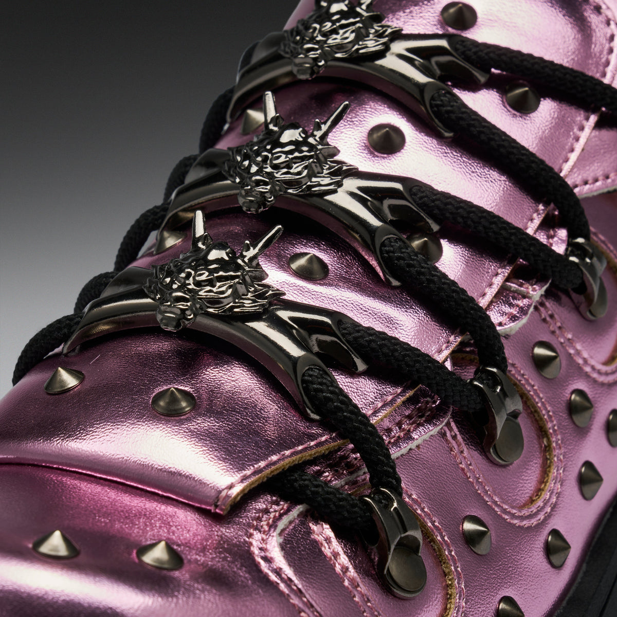 Close-up of a metallic pink high-top sneaker with black laces, silver conical studs, and ornate silver lace hardware featuring a wolf head design.