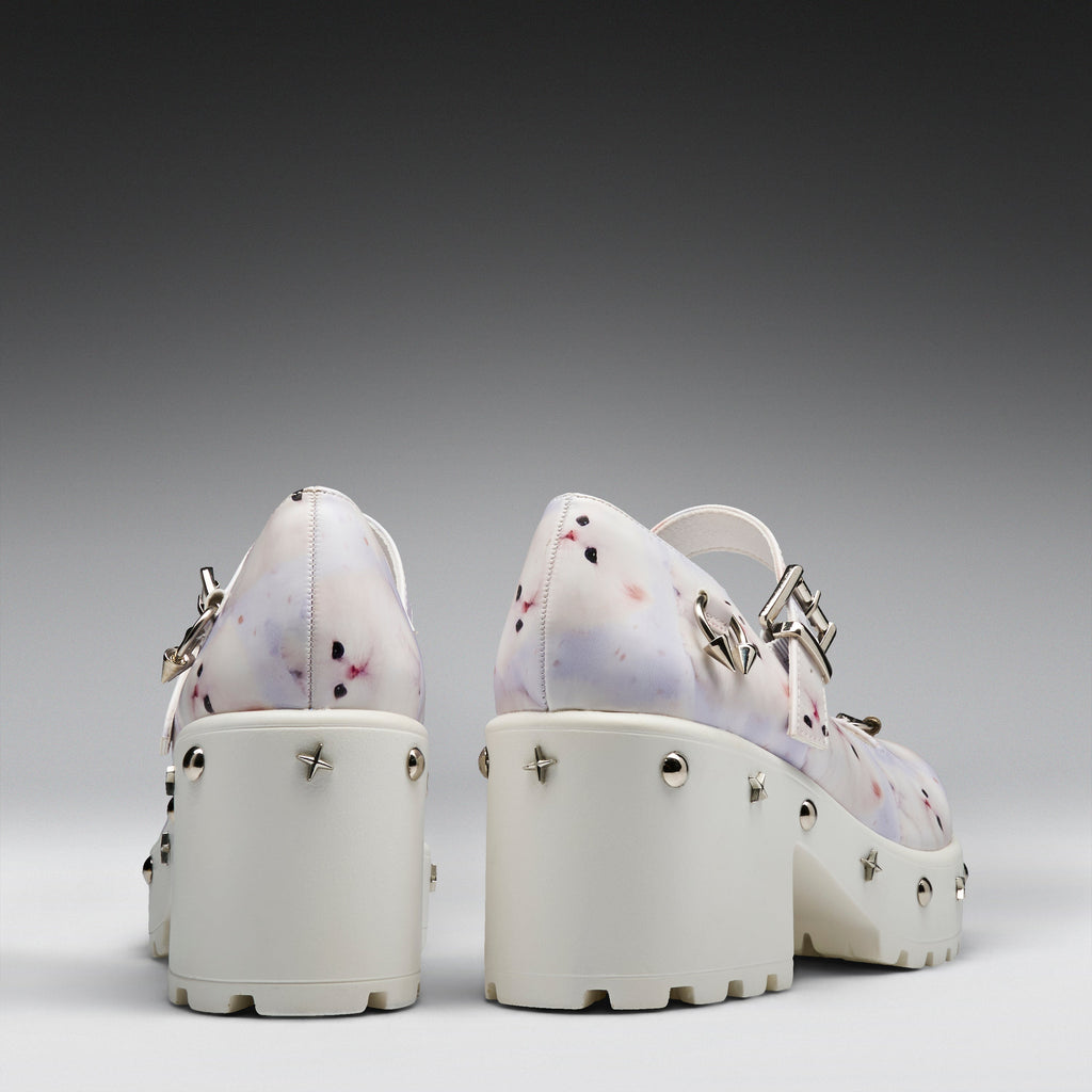 A pair of chunky platform Mary Jane shoes shown from the back, featuring a white base with a light purple and pink floral pattern, silver-tone metal studs, and buckle straps.