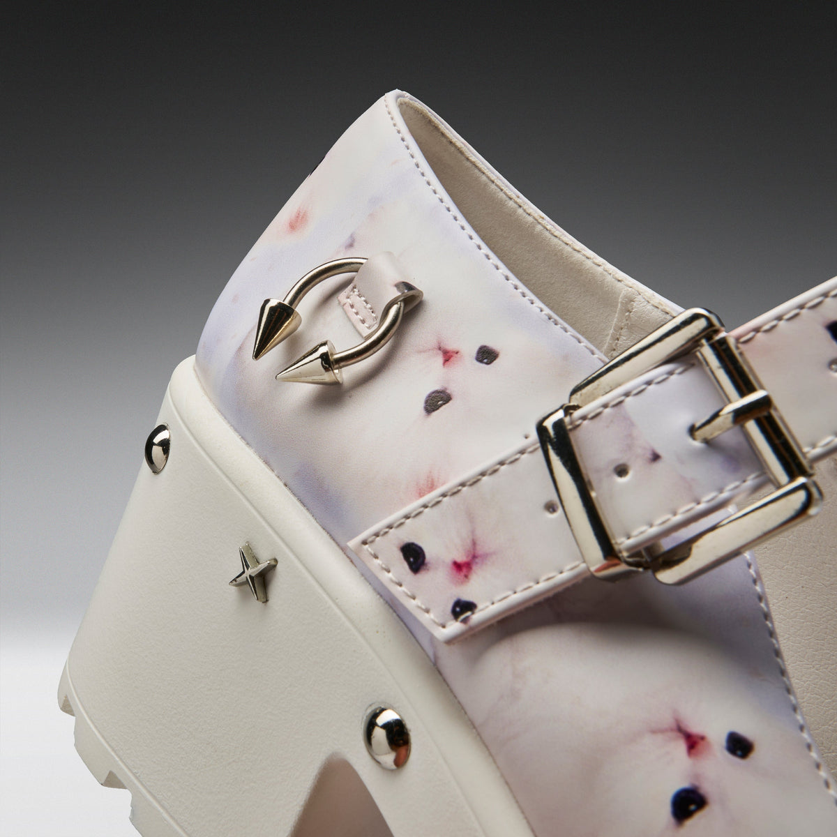 A close-up of a platform shoe featuring a glossy white and pale pink animal print with pink and black spots, decorated with silver-tone metal studs, a double-arrow ring, and a buckle strap.