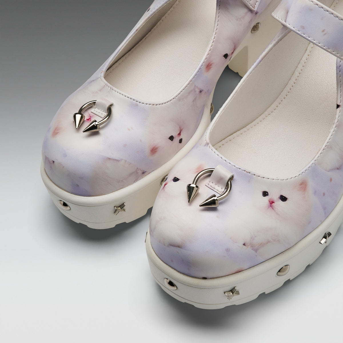 A pair of platform Mary Jane shoes featuring a pastel white and lavender cat print, decorated with silver metal hoop rings on the toes and small metallic studs on the soles.