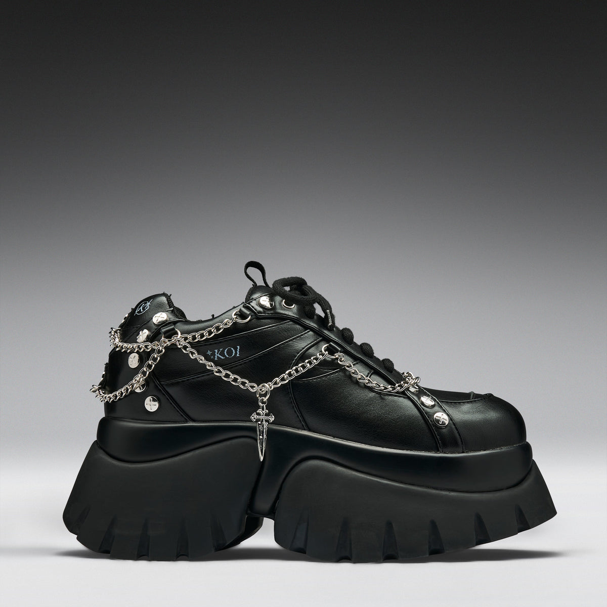 A black leather platform sneaker with chunky lug soles, silver chain details draped across the side, metal studs, and a small cross charm, shown against a dark gray gradient background.