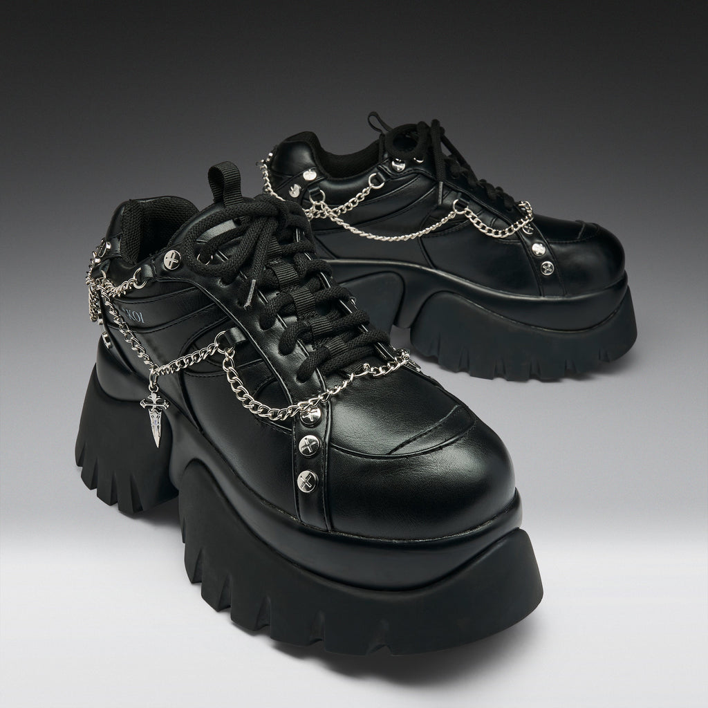 A pair of black leather platform sneakers with chunky lug soles, black laces, and silver chain details draped across the sides, photographed on a dark-to-light gray gradient studio background.