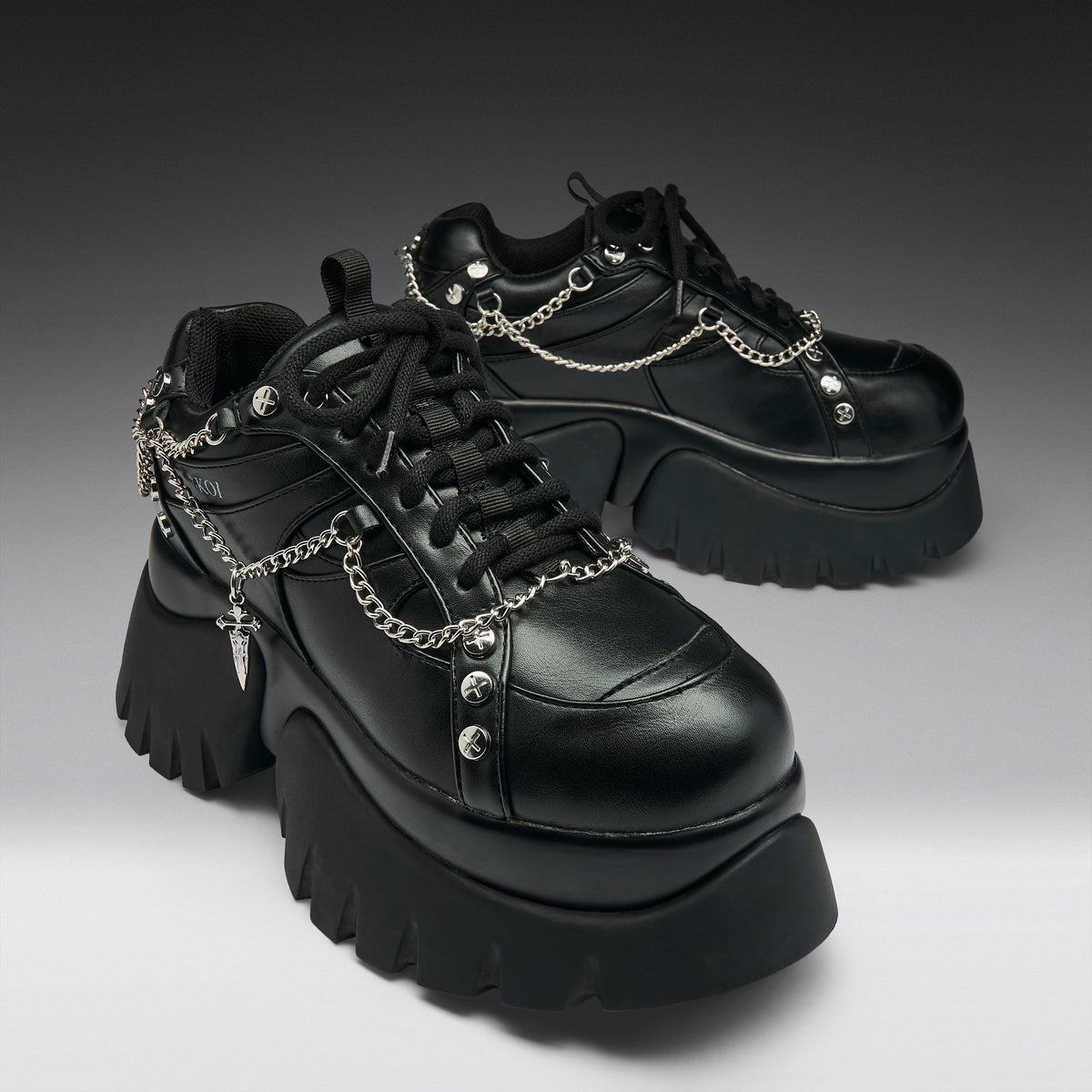 A pair of black leather platform sneakers with chunky lug soles, black laces, and silver chain details draped across the sides, photographed on a dark-to-light gray gradient studio background.