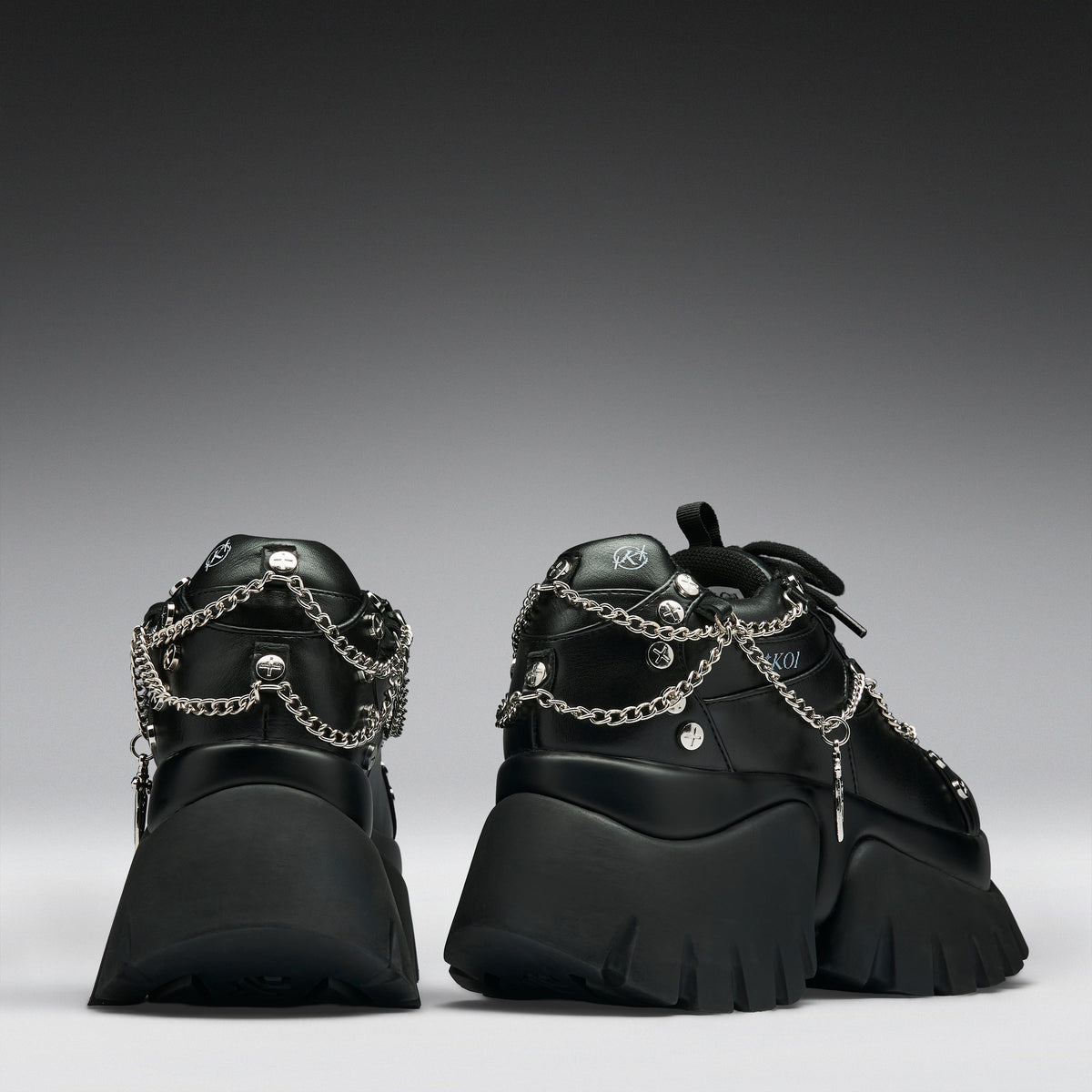 A pair of black chunky platform sneakers with silver chain and stud embellishments, shown from the back against a dark gray gradient background.