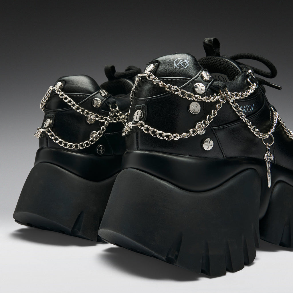 A pair of chunky black platform sneakers with silver chain straps, studs, and a small hanging cross charm, shown on a dark gray gradient studio background.