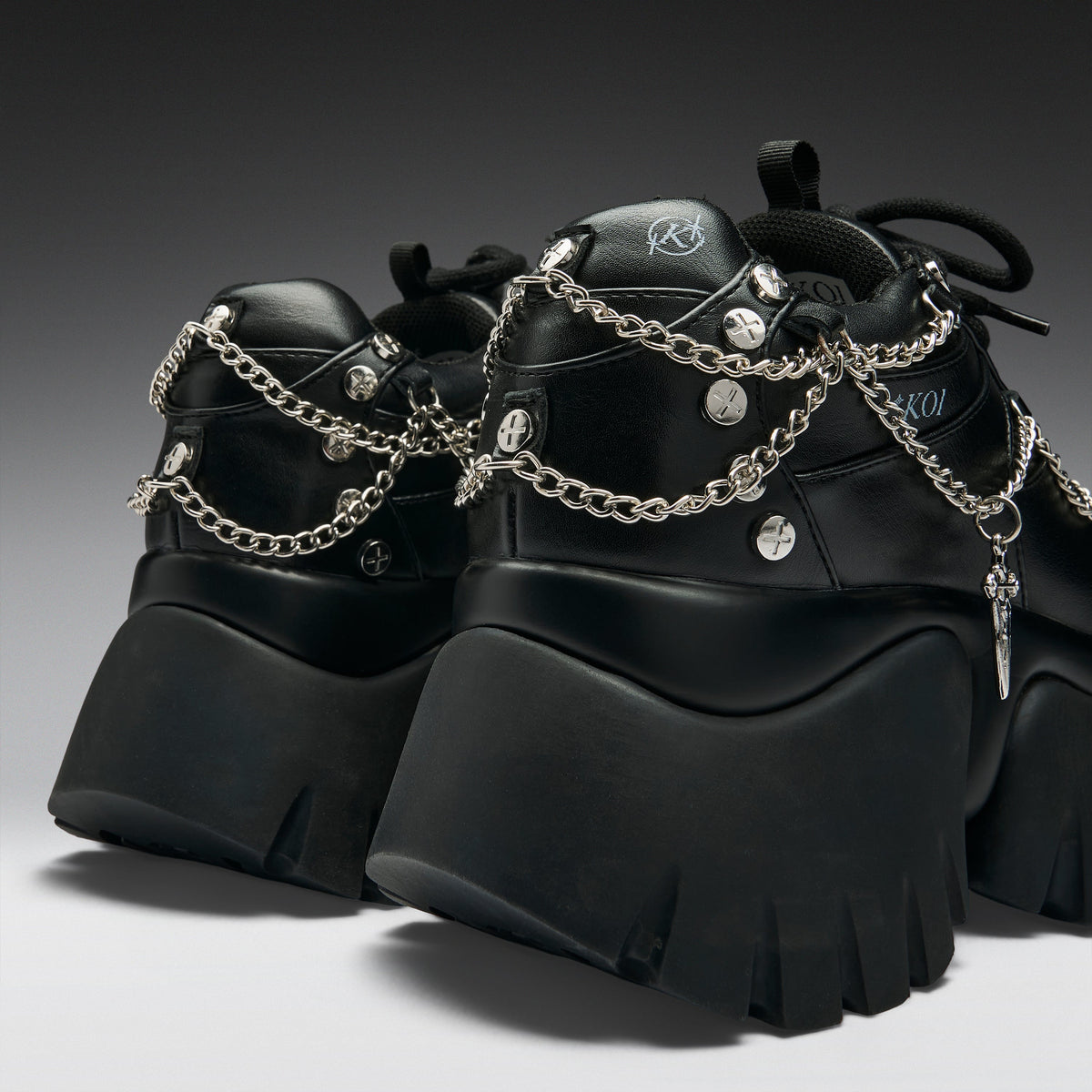 A pair of chunky black platform sneakers with silver chain straps, studs, and a small hanging cross charm, shown on a dark gray gradient studio background.