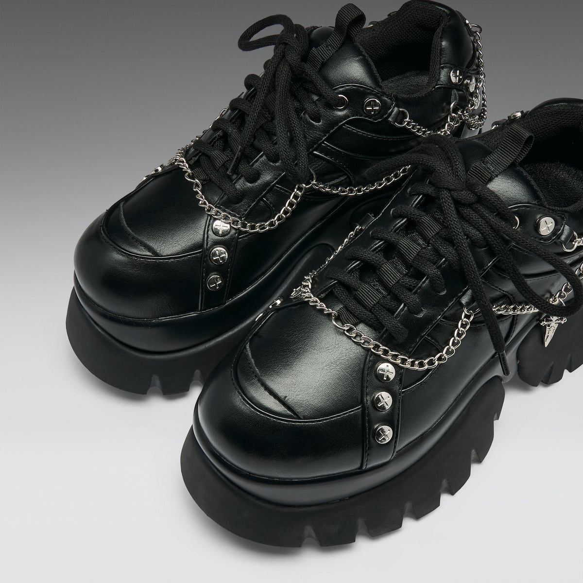 A pair of black leather platform sneakers with chunky lug soles, black laces, and silver chain details with skull and spike embellishments, shown on a gray gradient background.
