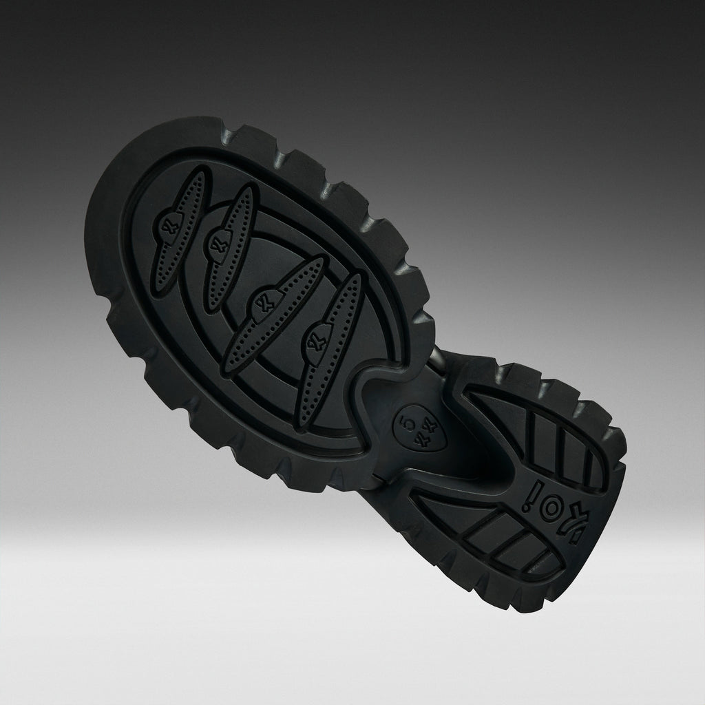 A close-up of a black rubber shoe sole with textured tread patterns and embossed branding, shown against a dark-to-light gray gradient background.