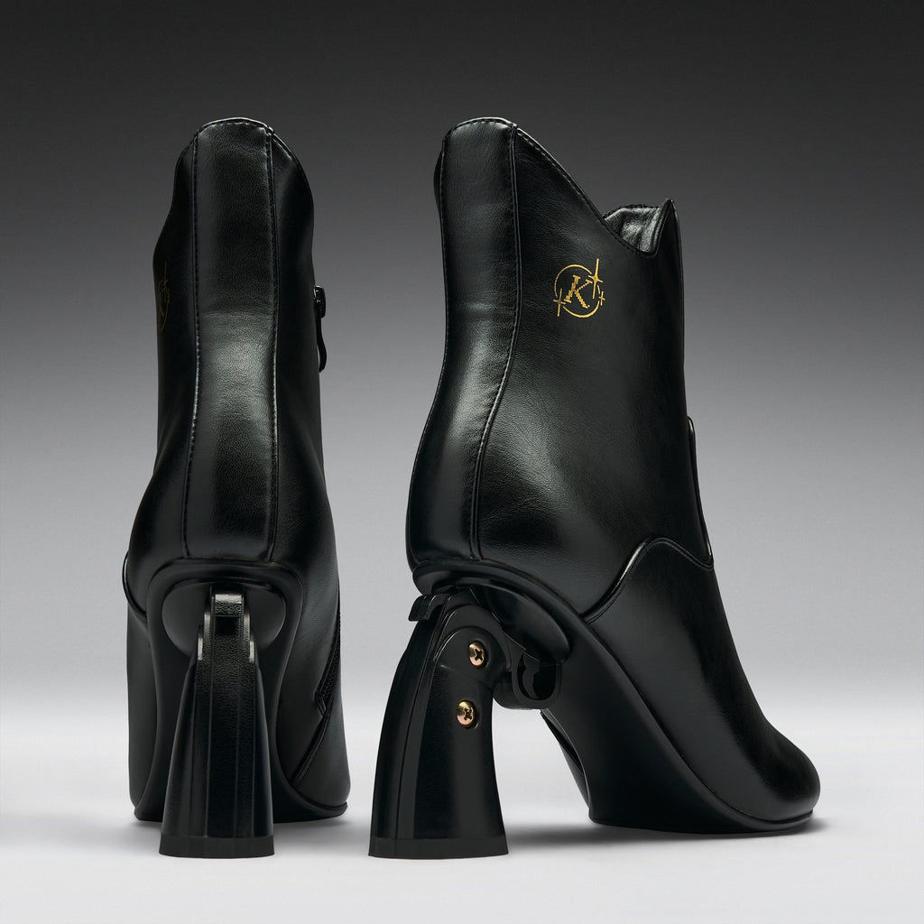 A pair of black leather ankle boots with chunky block heels, shown from the back and side against a dark gray gradient background. One boot features a gold circular 'RK' logo on the side.