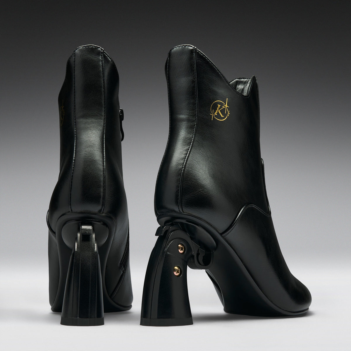 A pair of black leather ankle boots with chunky block heels, shown from the back and side against a dark gray gradient background. One boot features a gold circular 'RK' logo on the side.