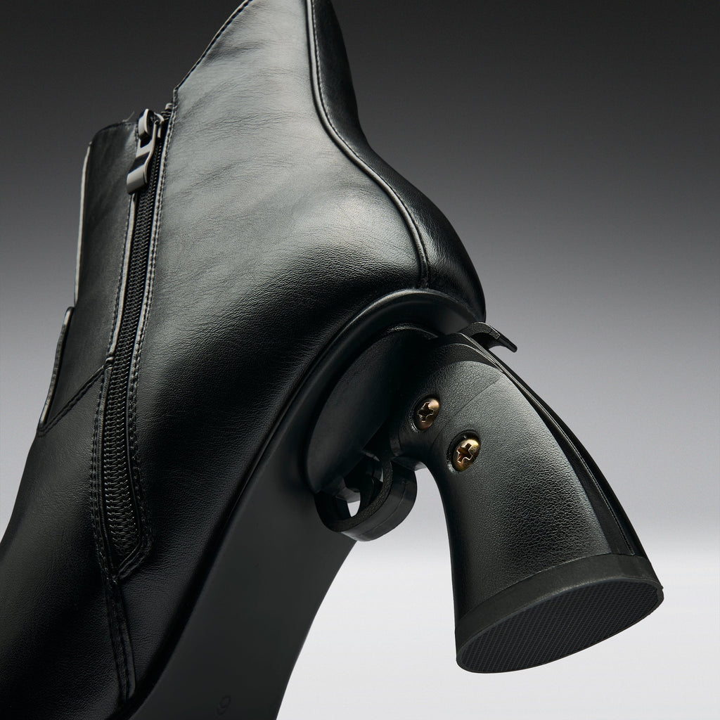 Close-up studio shot of a black leather ankle boot with a unique pistol-shaped heel and small gold-tone rivets, set against a dark gray gradient background.