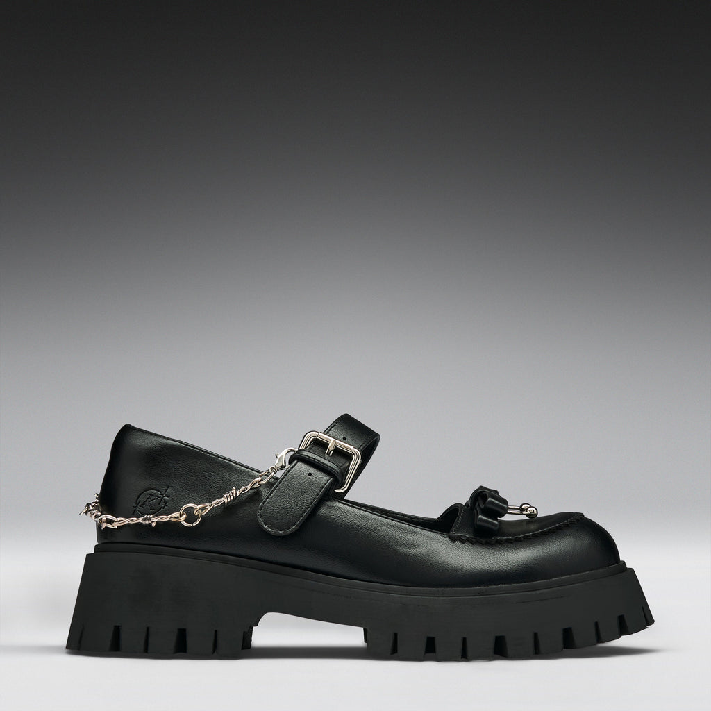 A black leather Mary Jane platform shoe with a chunky lug sole, a decorative tassel on the toe, and a silver chain draped from the buckle, shown against a dark gray gradient background.