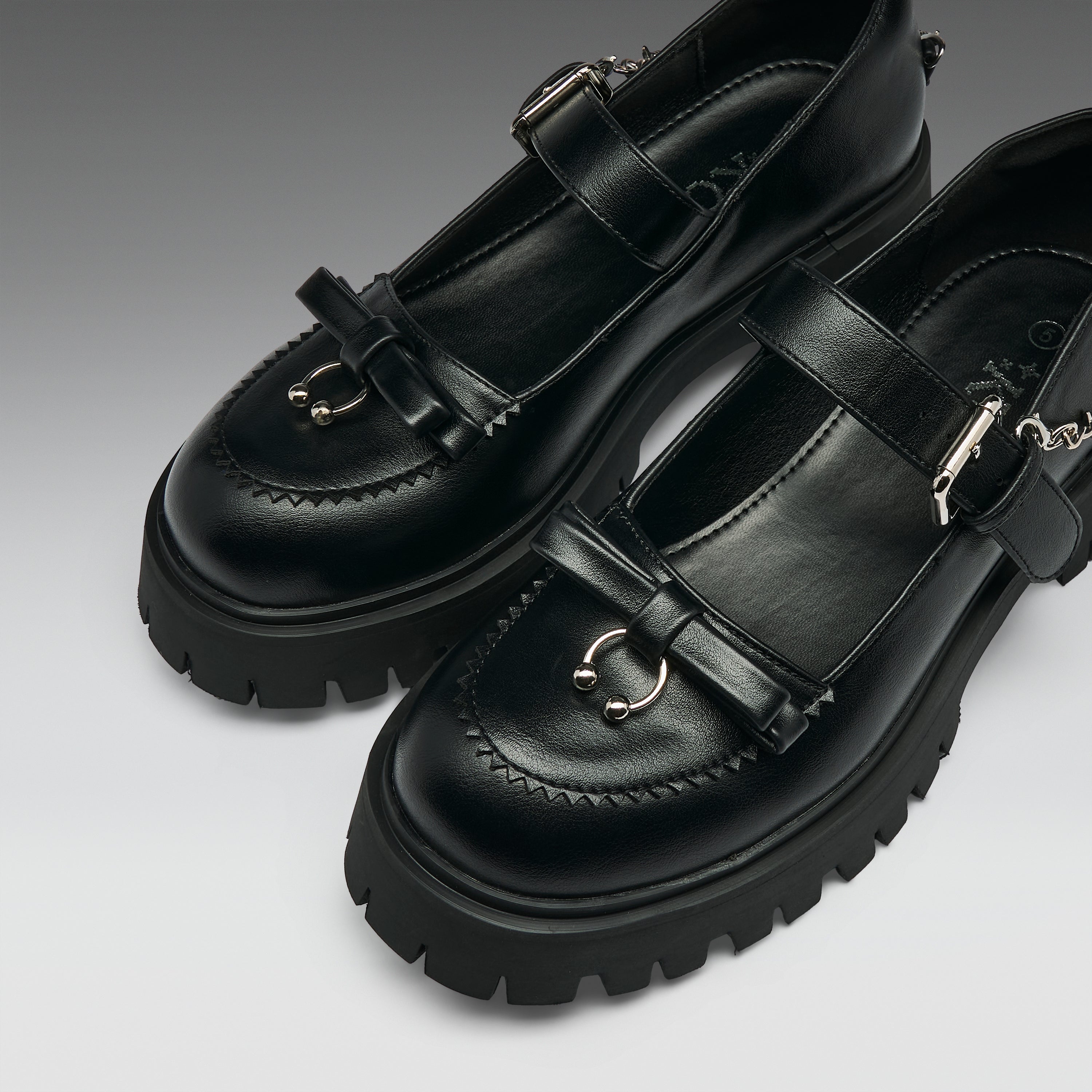 A pair of black leather Mary Jane-style shoes with chunky lug soles, decorative bow details, and silver-tone metal hardware, photographed on a clean gray studio background.